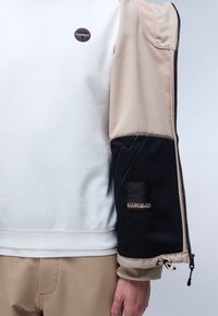 Vit sweatshirt med beige och svarta texturerade ärmar. Har en logotyp patch på bröstet och ett dragkedjedetalj på ärmen. Slätt tyg med accenter.