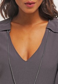 Graue Bluse mit V-Ausschnitt, strukturierter Oberfläche und Kragen-Detail. Eine silberne Kettenhalskette ist sichtbar und betont den Ausschnitt.