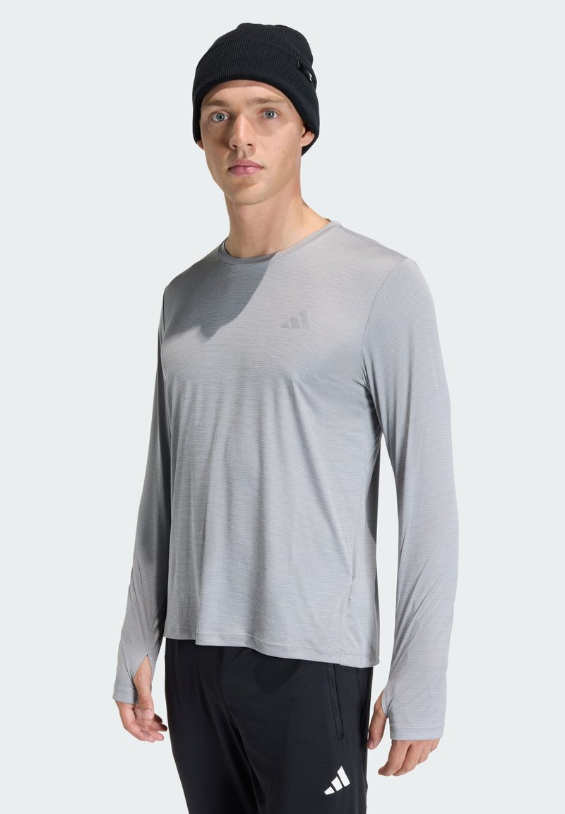 Light grey long-sleeve athletic shirt with round neck and side slits. Black beanie worn. Features subtle striped texture and logo.
