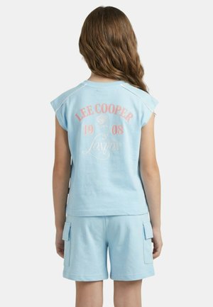 Enfant aux cheveux longs portant un haut sans manches bleu clair et un short assorti avec des poches latérales, debout, de dos à la caméra.