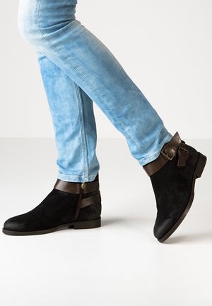Sorte ruskindsankelstøvler med brune læderstropper, med lynlås i siden og flad sål, parret med lyseblå denim jeans.