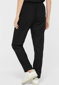 Person, der schwarze Cordhosen und weiße Veja-Sneaker trägt, steht mit einem Bein hinter dem anderen auf einem einfarbigen Hintergrund.