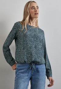 Blusa a maniche lunghe di colore verde scuro con un motivo testurizzato che presenta macchie blu chiaro e bianche, rifinita con un fiocco all'orlo. Abbinata a jeans blu.