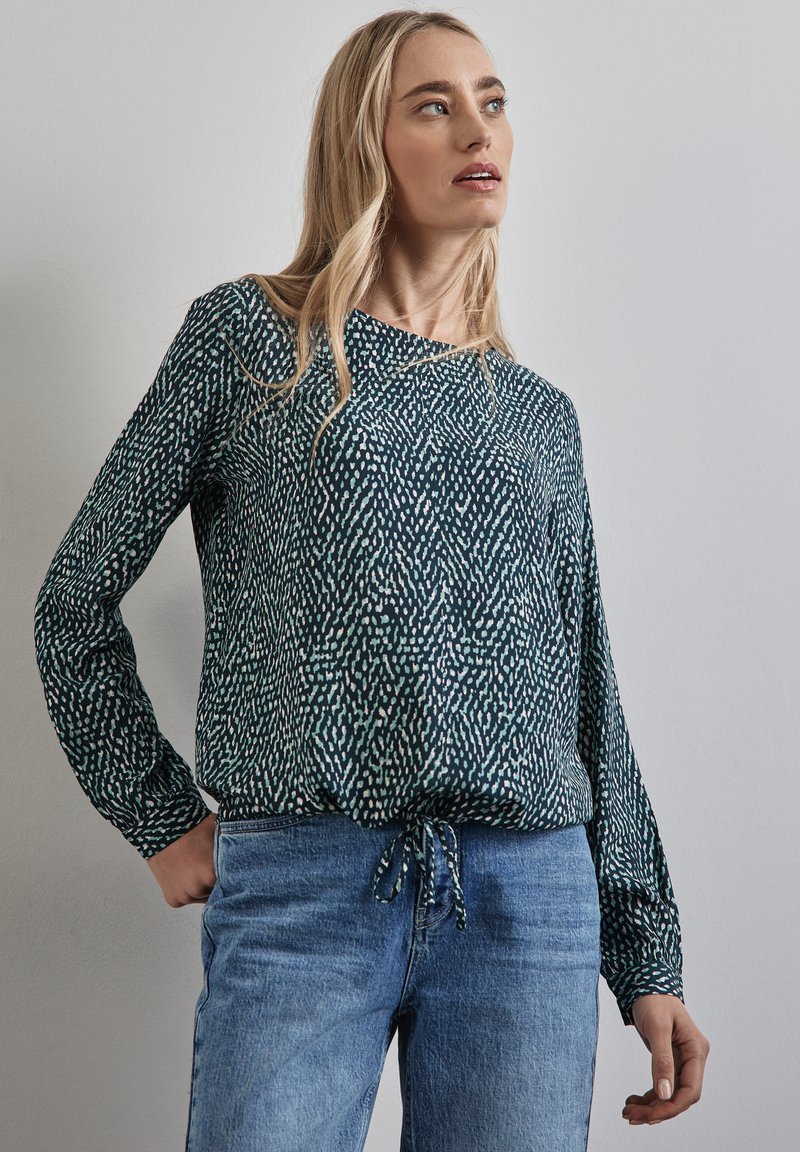 Blusa a maniche lunghe di colore verde scuro con un motivo testurizzato che presenta macchie blu chiaro e bianche, rifinita con un fiocco all'orlo. Abbinata a jeans blu.