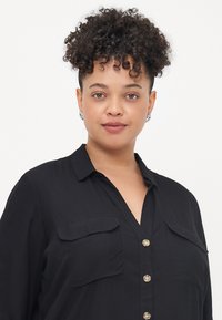 Chemise noire à boutons avec deux poches poitrine, col et boutons en ton doré. Tissu lisse avec une coupe décontractée, adaptée pour un style décontracté.