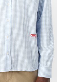Light blue and white checked long-sleeve shirt with button front, cuffs, and red "POMPE" logo. Made from woven fabric, casual fit.