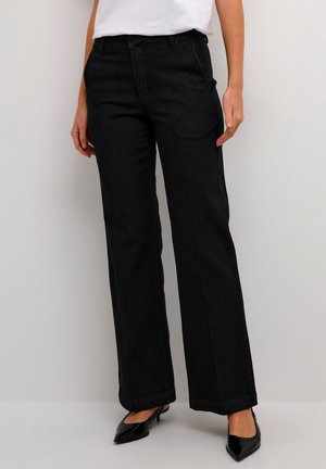 Femme portant un jean noir droit, un haut blanc et des talons noirs à bout pointu, debout devant un fond clair uni.