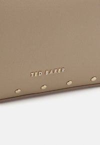 Portafoglio in pelle beige con borchie in metallo e logo "TED BAKER" impresso. Texture liscia, forma rettangolare, design minimalista.
