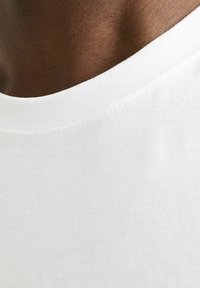 Hvid bomulds T-shirt krave, ribbede struktur, nærbillede af udskæringen mod mørkere hud, glat sømdetalje synlig.