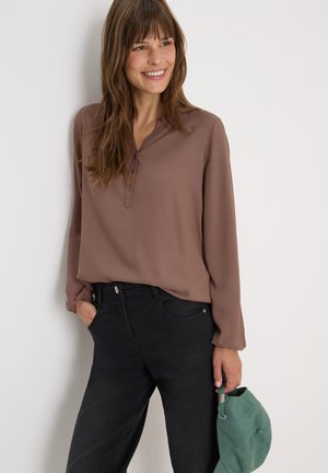 Blouse marron à manches longues en tissu léger avec un col henley et des boutons, associée à un jean noir et portant une casquette verte.