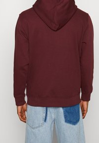 Burgundowa bluza z kapturem ma miękką teksturę, długie rękawy i ściągacz u dołu. Model nosi ją z jasnoniebieskimi dżinsami z naszywkami.