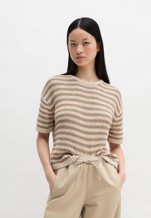Młoda kobieta z długimi czarnymi włosami, ubrana w beżowo-białą, paskowaną, dzianinową bluzkę z krótkim rękawem oraz beżowe spodnie ze ściągaczem, z rękami w kieszeniach.