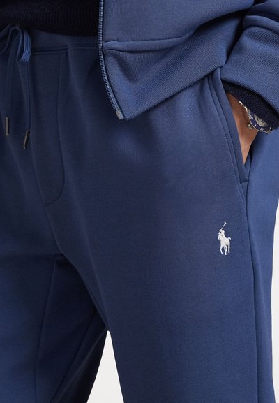 Σκούρες μπλέ φόρμες από μαλακό υλικό, με λευκό λογότυπο στο μηρό, ελαστική μέση και πλαϊνές τσέπες με διακριτική ραφή.