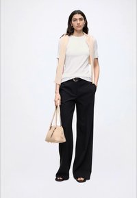 Femme debout portant une chemise blanche à manches courtes, un pull sans manches beige drapé sur les épaules, un pantalon noir à jambes larges, et tenant un sac à main beige.