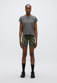 Γκρι κοντομάνικη μπλούζα σε συνδυασμό με πράσινα σορτς καμουφλάζ. Το μοντέλο φορά μαύρες κάλτσες μέχρι το γόνατο και μαύρα αθλητικά παπούτσια. Απλός, λειτουργικός σχεδιασμός.