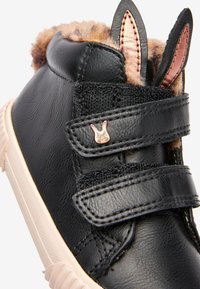 Schwarzer Kunstleder-Schuh mit rosa Akzenten, ausgestattet mit Hasenohren-Details, einem flauschigen Futter und doppelten Klettverschlüssen.