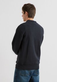 Marineblauwe gebreide trui met een textuurpatroon, geribde manchetten en zoom. Gecombineerd met blauwe spijkerbroek, gezien vanuit de achterkant, met de kraag in het middelpunt.