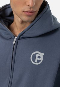 Plavajak s kapuljačom koji se zatvara na patentni zatvarač, s srebrnim logotipom na prsima. Glatka tekstura s vidljivim detaljem patentnog zatvarača i kapuljače.