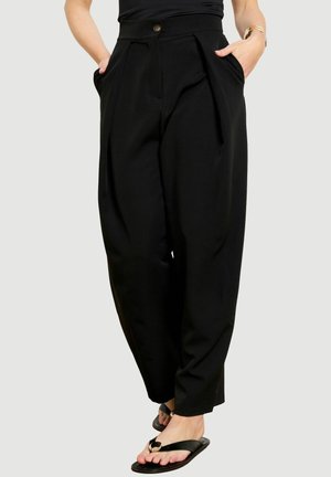 Frau trägt hoch taillierte schwarze schmale Hose mit vorderen Falten und Taschen, kombiniert mit schwarzen flachen Sandalen und einem schwarzen Oberteil.