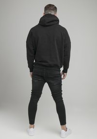 Felpa nera con cappuccio ampio, vestibilità oversize e polsini a costine, abbinata a jeans scuri e sneaker bianche, vista da dietro.