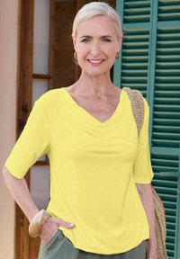 Gelb drapierte Bluse mit kurzen Ärmeln, glatter Textur, lockere Passform, kombiniert mit grünen Hosen und einer gewebten Tragetasche.