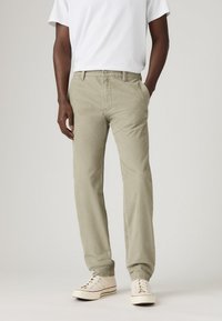 Man wearing beige corduroy pants, white sneakers, and a white short-sleeve shirt, standing with one hand in pocket on a plain background.