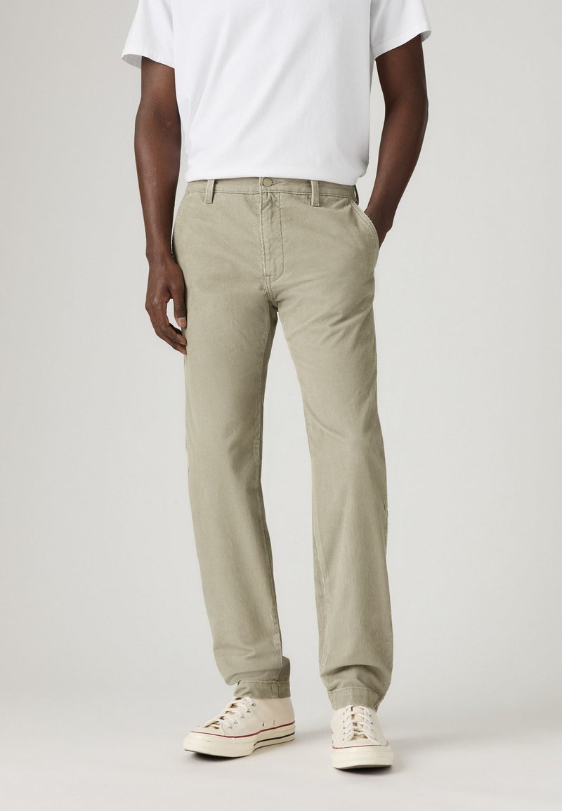 Man wearing beige corduroy pants, white sneakers, and a white short-sleeve shirt, standing with one hand in pocket on a plain background.
