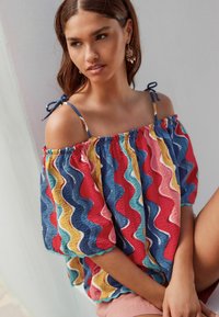 Haut coloré à décolleté asymétrique présentant un motif ondulé en rouge, bleu, jaune et rose ; tissu léger avec des manches courtes et des bords festonnés décoratifs.