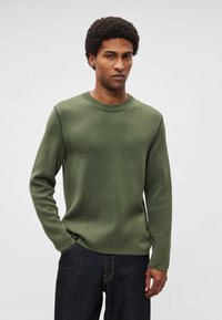 Camisola verde de malha com decote redondo, mangas compridas e costuras em contraste ao longo das ombreiras. Usada com jeans de ganga escura.