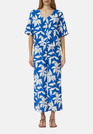 Frau trägt blaues Kurzarm-Oberteil und weit geschnittene Hosen mit großem weißem tropischem Blattmuster, kombiniert mit braunen Sandalen.