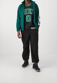 Giacca sportiva verde con zip, canotta NBA nera con "Celtics" e numero "0", pantaloni neri larghi e sneakers nere con dettagli viola.
