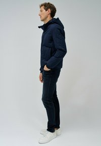 Chaqueta acolchada de color navy con capucha, que presenta un cierre de cremallera y bolsillos frontales. Combinada con jeans oscuros y zapatillas blancas. Diseño simple y moderno.