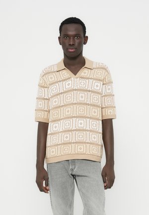 Pletená béžová polokošeľa s výstrihom do V, zdobená bielymi geometrickými vzormi a krátkymi rukávmi. Mäkká textúra, ľahký dizajn.