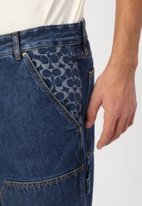 Jeans denim w ciemnoniebieskim odcieniu, z wyrazistym wzorem na kieszeniach. Projekt zawiera subtelne szwy oraz prosty krój.