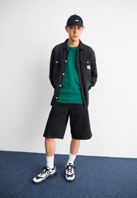 Svart skjorta med knappar över en grön t-shirt, svarta shorts, vita strumpor och svart-vita sneakers. Golvet är blått heltäckningsmatta, bakgrunden är enkel.