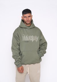 Felpa verde oliva con tasca frontale, vestibilità oversize e stampa grafica bianca "multiply". Abbinata a pantaloni della tuta beige con logo.