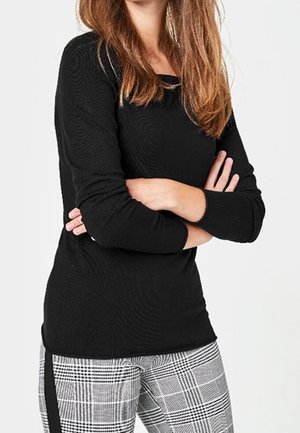 Femme aux longs cheveux bruns portant un haut noir à manches longues et un pantalon à carreaux noir et blanc, bras croisés sur la poitrine.