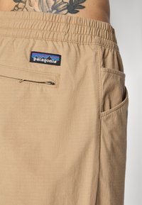 Beige Patagonia shorts with elastic waistband, rear zip pocket, side pocket, worn by a tattooed person with floral back tattoo visible.