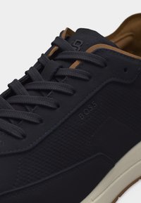 Σκούρο μπλε αθλητικό παπούτσι με υφασμάτινες πλευρές, κορδόνια στο ίδιο χρώμα, καφέ τρέσα, ανάγλυφο λογότυπο "BOSS" και λευκή σόλα.