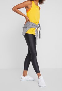 Keltainen urheilullinen toppi, jonka vyötärölle on sidottu harmaa neuletakki, yhdistetty mustiin leggings-housuihin ja valkoisiin tennareihin. Pehmeä kangas ja vartalonmyötäinen malli.