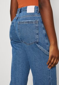 Höga blå denimjeans med rak skärning, med bakfickor, synlig sömnad och en rosa tygmärke på midjebandet.