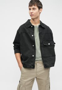 Chaqueta de denim negra con botones a presión y ajuste oversize, combinada con un suéter de cuello redondo de color verde claro y pantalones cargo caqui. Diseño minimalista.