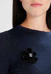 Schwarze Samt-Brosche mit schleifenartigem Design, versehen mit einem zentralen klaren Strassstein, befestigt an einem dunkelblauen Pullover.