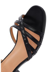 Zwarte sandaal met hakken, voorzien van kruislings straps, een gladde matte textuur en een nep slangenhuidpatroon op sommige straps. Lichte beige zool.
