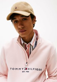 Felpa con zip color rosa chiaro con "TOMMY HILFIGER EST. 1985" in navy, abbinata a una camicia a righe multicolore e a un cappellino beige.