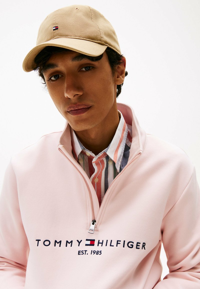 Felpa con zip color rosa chiaro con "TOMMY HILFIGER EST. 1985" in navy, abbinata a una camicia a righe multicolore e a un cappellino beige.