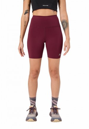 Personne portant un short de cycliste bordeaux taille haute, un crop top noir, des chaussettes grises à motifs, et des chaussures de course, debout les bras détendus.