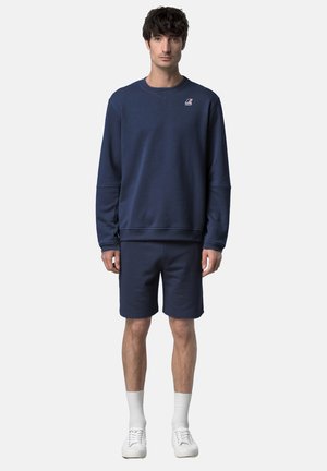 Uomo che indossa una felpa blu navy e pantaloncini abbinati blu navy, calzini bianchi e scarpe da ginnastica bianche, in piedi davanti a uno sfondo chiaro e uniforme.