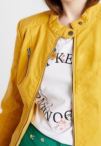 Giacca di pelle gialla con colletto e chiusura con zip, indossata sopra una camicia bianca con ricami floreali e testo nero. Pantaloni verdi visibili.