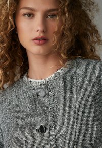 Šedá tkaná bunda s texturovaným povrchem, vybavená třásňovým límcem a dvěma kulatými černými knoflíky vpředu. Jednoduchý, čistý design.